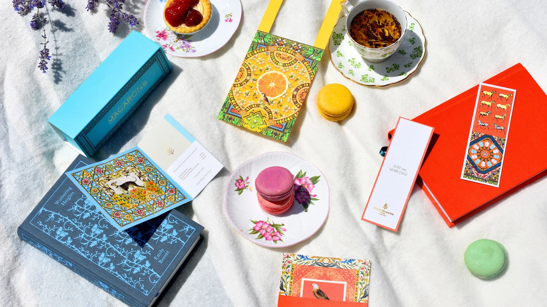 Styled flat lay of Four Seasons branded items including illustrated cards, gift boxes, tea cups, macarons, and printed collateral arranged on a light fabric background.