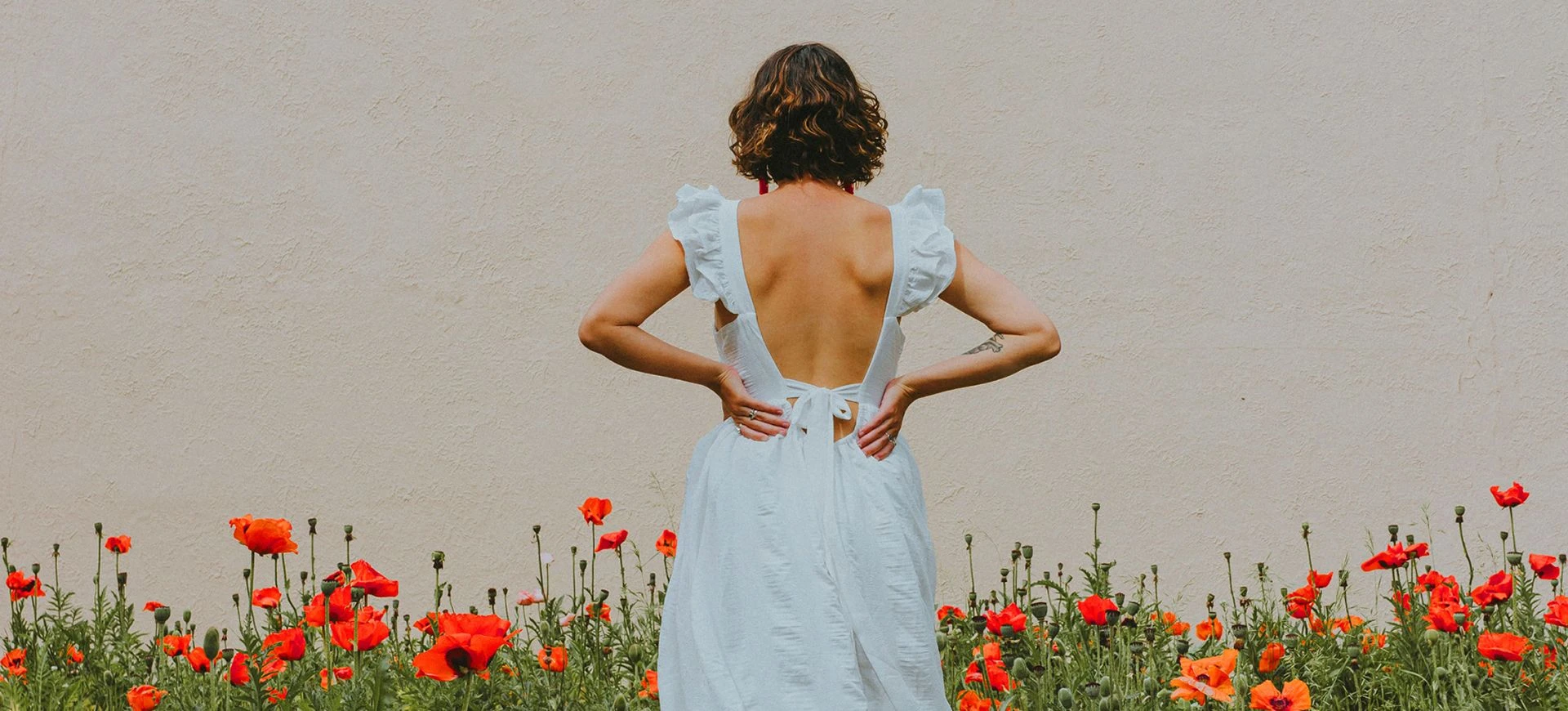 Person standing with their back to the camera wearing a white open-back dress, surrounded by red flowers against a neutral wall.