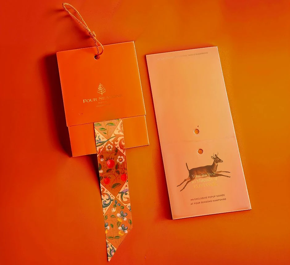 Twilly invitation featuring a patterned silk ribbon attached to an orange invite card with gold foil Four Seasons branding.